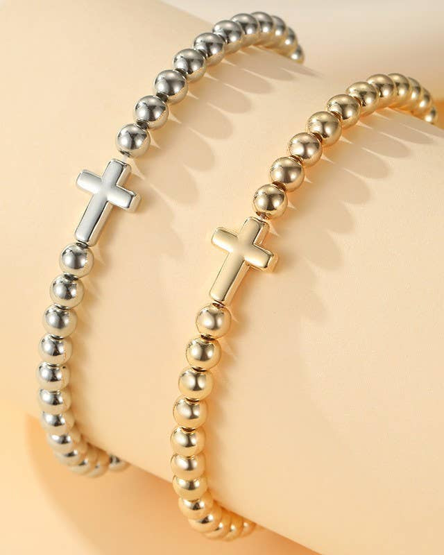 Two beaded stretch bracelets with cross charms, one in silver and one in gold, displayed on a beige jewelry post.