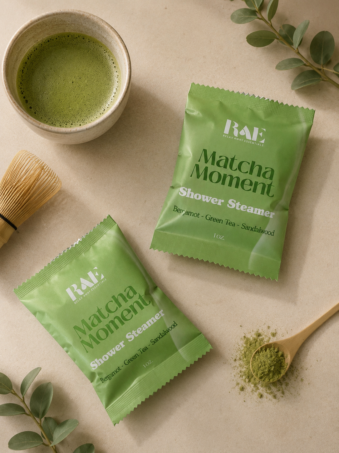 RAE Matcha Moment shower steamer packets with a cup of matcha, wooden spoon, and green leaves on a beige background.