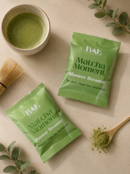 RAE Matcha Moment shower steamer packets with a cup of matcha, wooden spoon, and green leaves on a beige background.