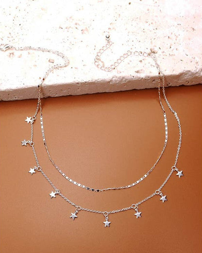 A double-strand silver necklace with small star-shaped charms, displayed on a textured terracotta surface with a light stone slab at the top.