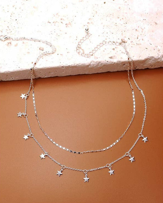 A double-strand silver necklace with small star-shaped charms, displayed on a textured terracotta surface with a light stone slab at the top.