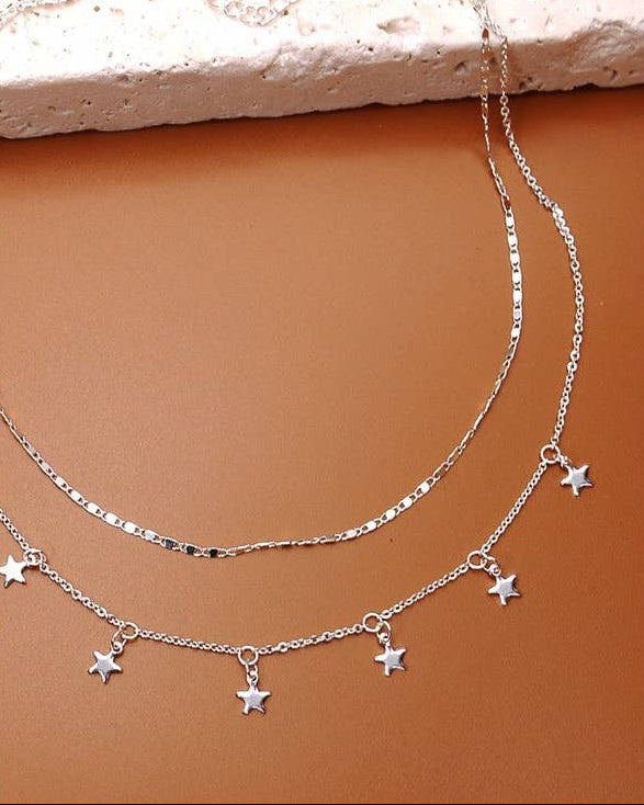 A layered silver necklace set displayed on a warm brown surface, featuring a longer chain and a shorter chain with five dangling star charms.