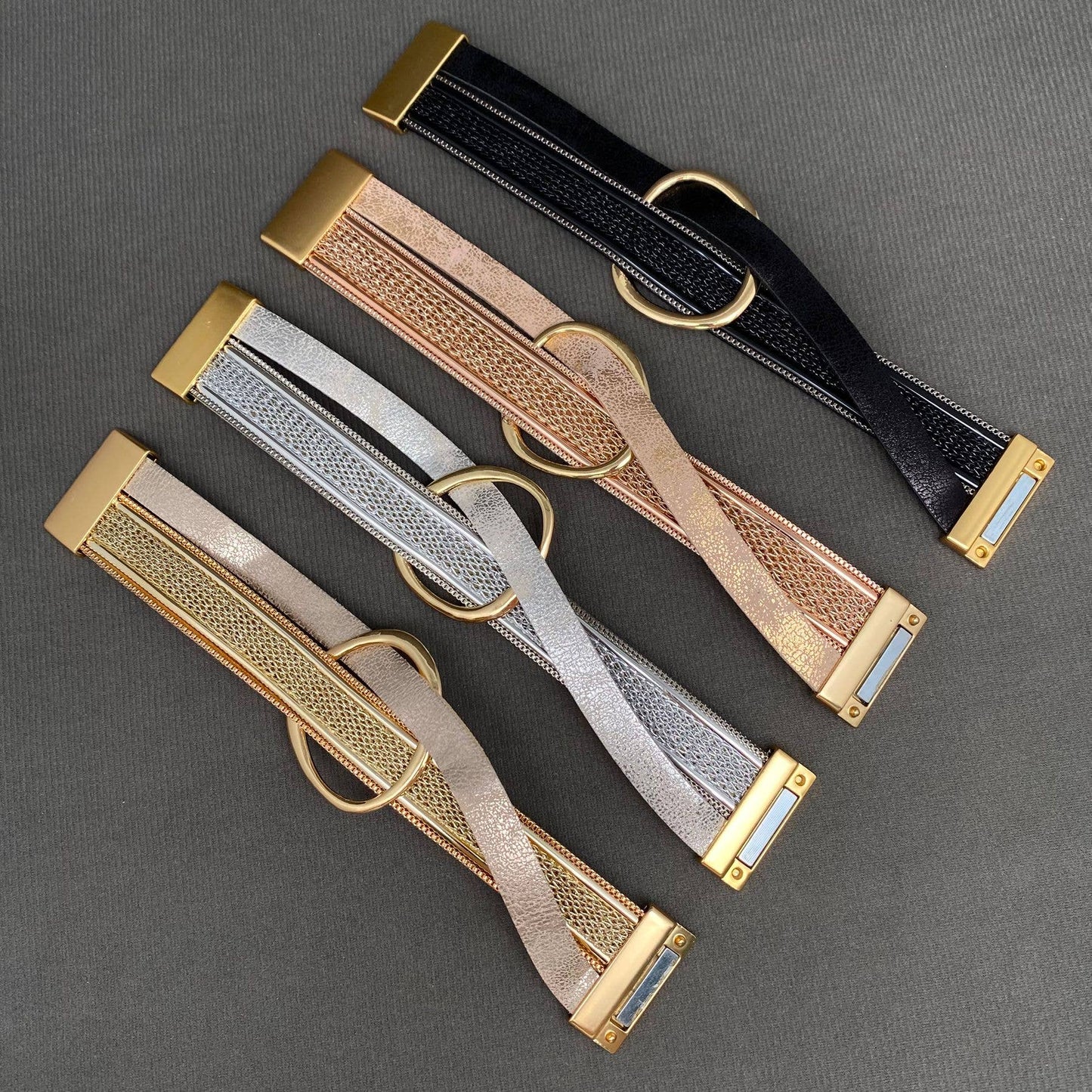 Four double wrap woven leather bracelets with gold-tone hardware, arranged diagonally on a dark gray textured background. The four shown are in black, rose gold, silver, and champagne gold.