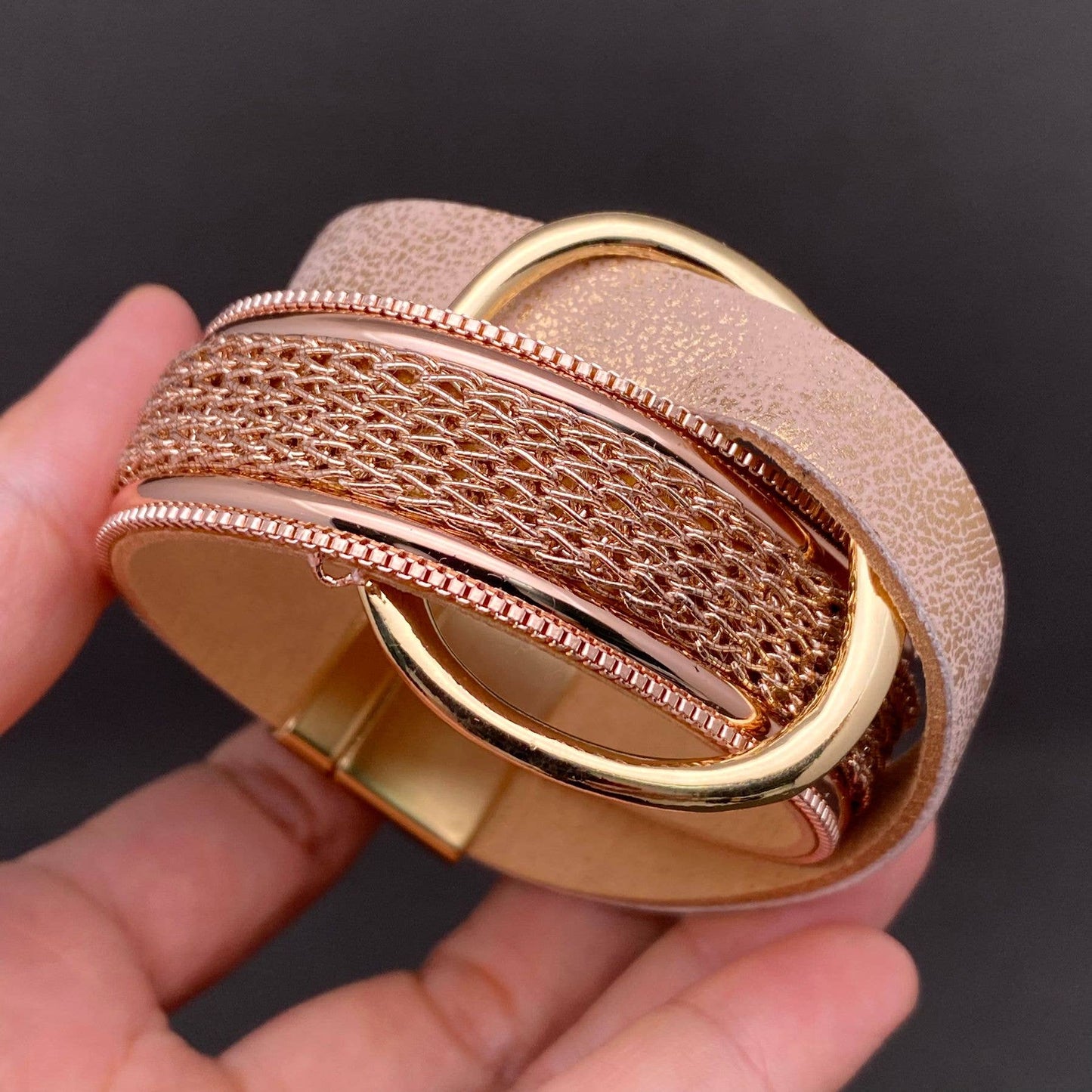 A close-up of a chunky leather woven cuff bracelet held in a hand. The bracelet features a textured light rose gold woven band layered with metallic rose gold mesh and polished gold-tone rings.
