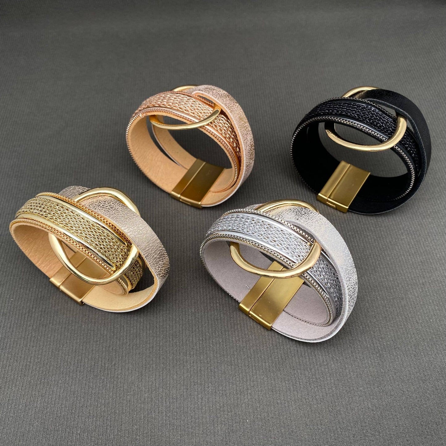 Four wide woven leather bracelets with large oval gold-tone rings, shown in rose gold, black, silver, and gold on a dark gray textured background.