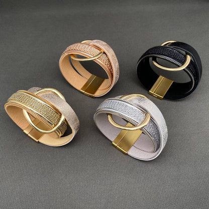 Four wide woven leather bracelets with large oval gold-tone rings, shown in rose gold, black, silver, and gold on a dark gray textured background.