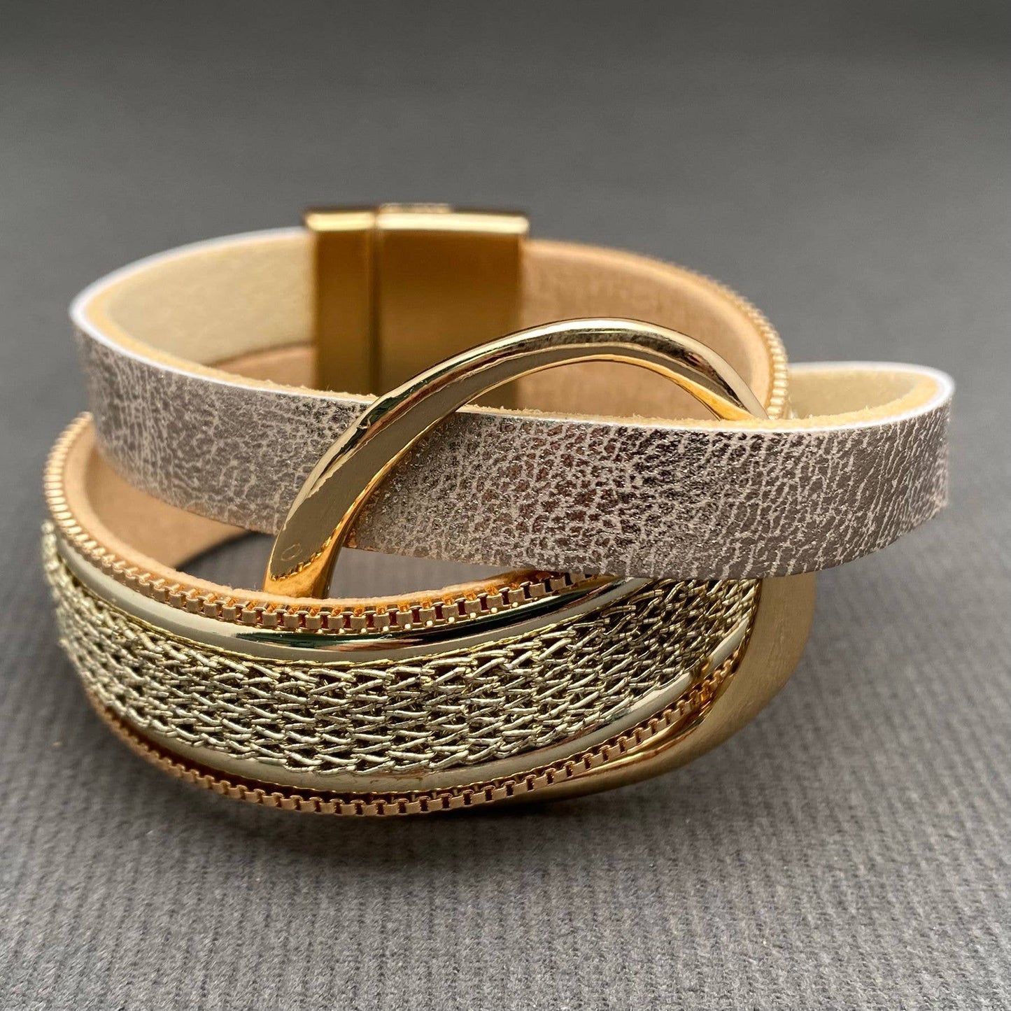 A multi-layered leather and metal cuff bracelet featuring a textured light beige leather band, a gold-tone chain link section, and polished gold-tone metal ring, displayed on a gray textured background.