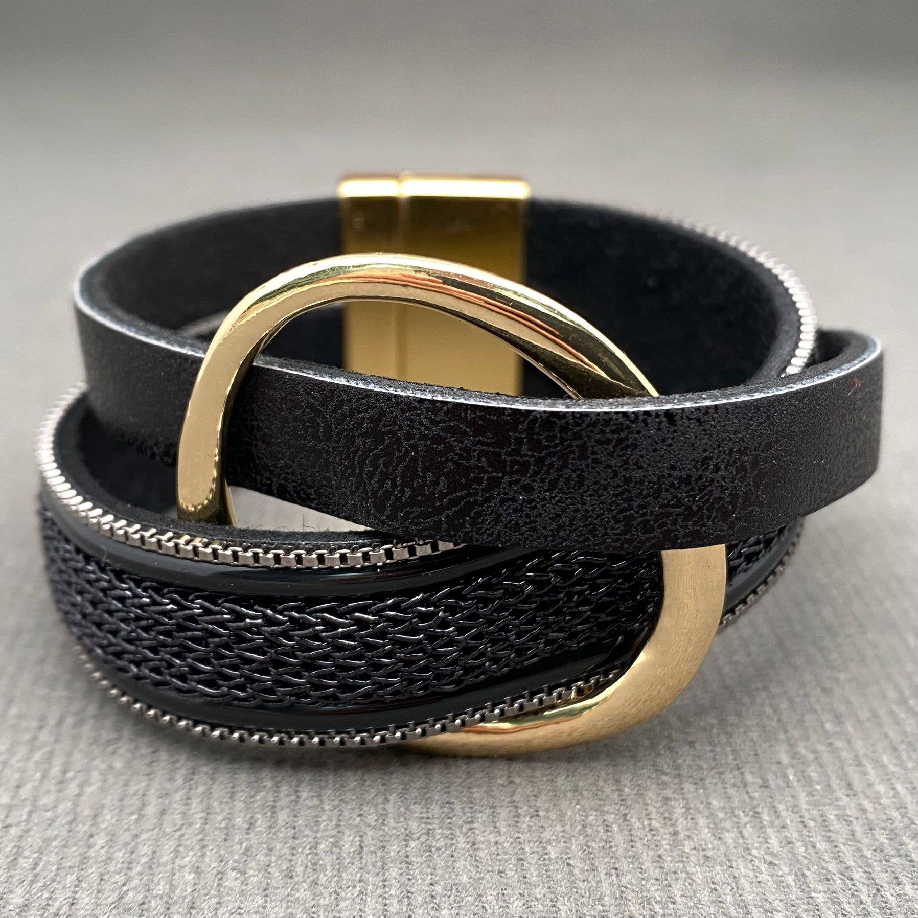 A black leather wrap bracelet with a textured woven center stripe, a gold-tone metal ring, and a gold-tone magnetic clasp, displayed on a neutral gray textured surface.