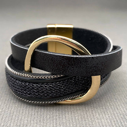 A black leather wrap bracelet with a textured woven center stripe, a gold-tone metal ring, and a gold-tone magnetic clasp, displayed on a neutral gray textured surface.