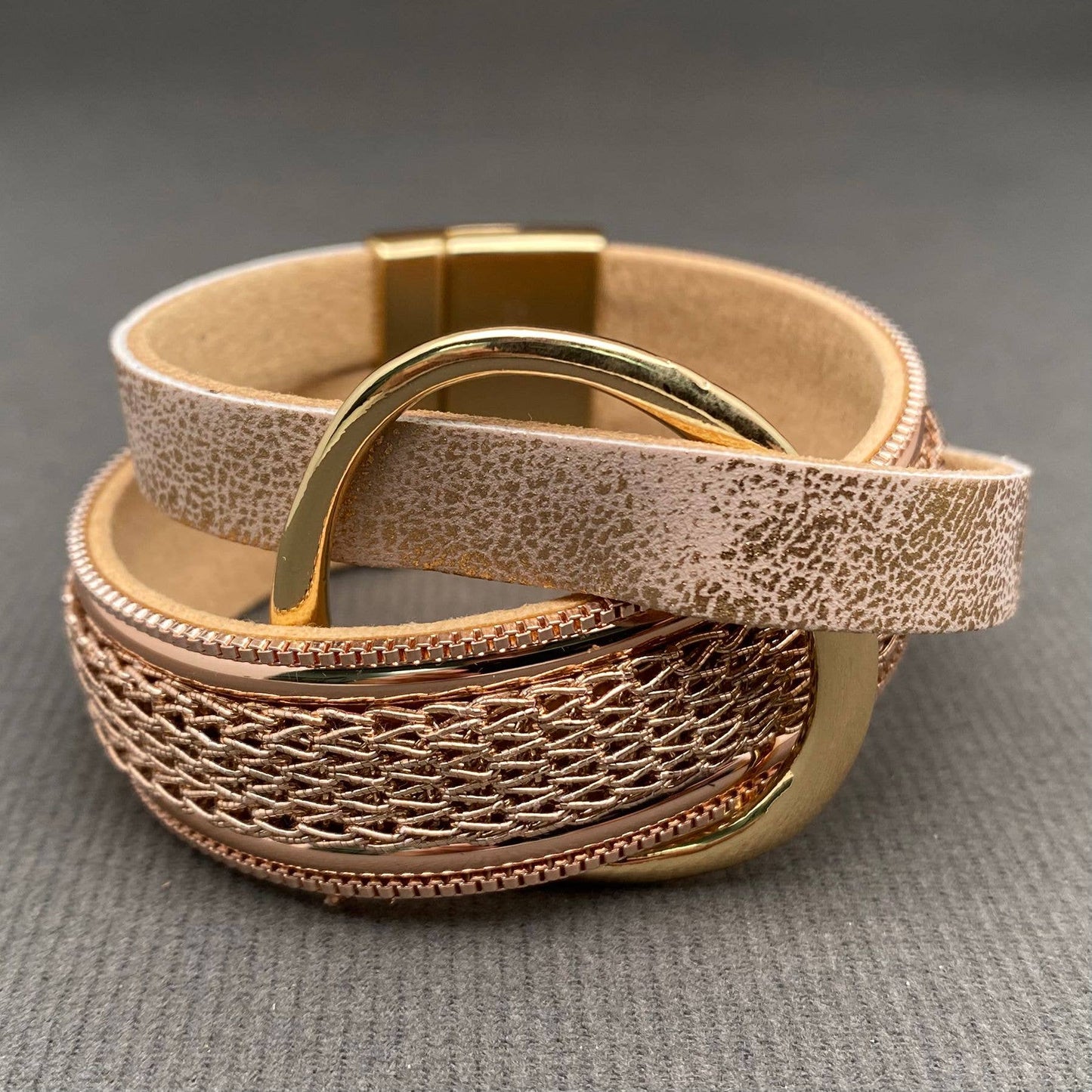 A close-up of a woven leather bracelet with a textured beige exterior, a metallic rose gold center band, and polished gold-toned hardware on a gray textured surface.