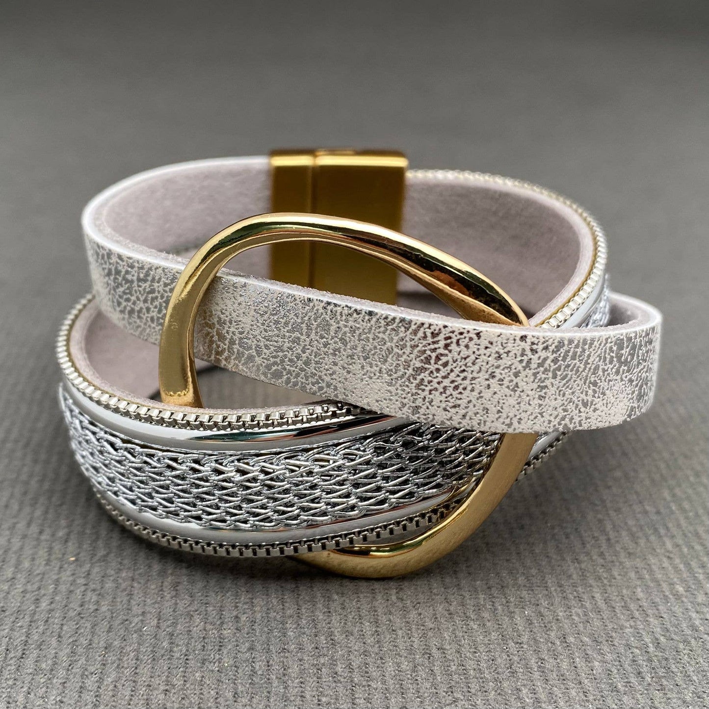A multi-layered leather bracelet with textured silver layers, a woven silver center band, and polished gold-tone metal hardware, including a large ring and clasp, displayed on a gray textured background.