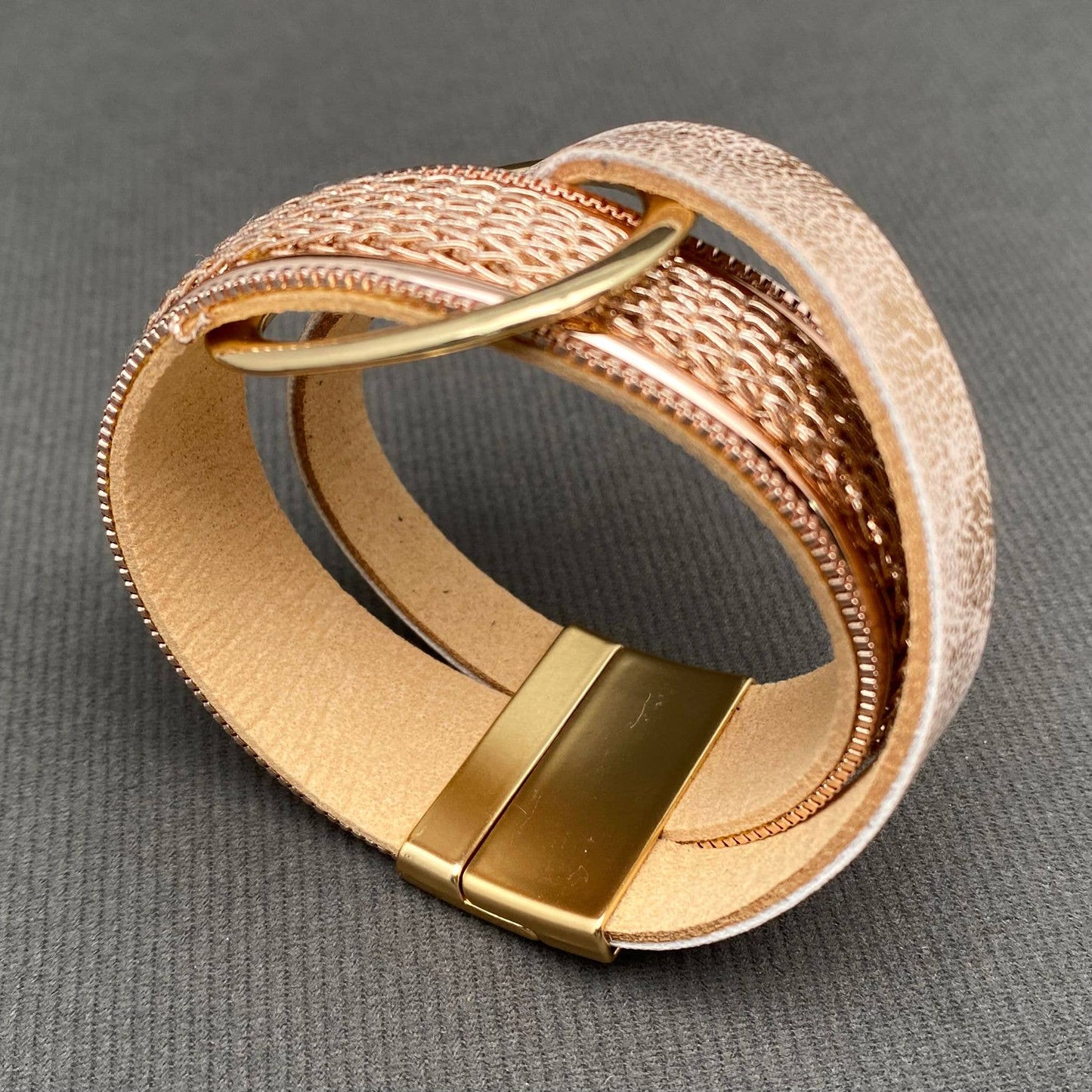 A wide cuff bracelet made of gold-toned metal and rose gold leather, featuring a polished gold-toned clasp, photographed on a textured gray background.