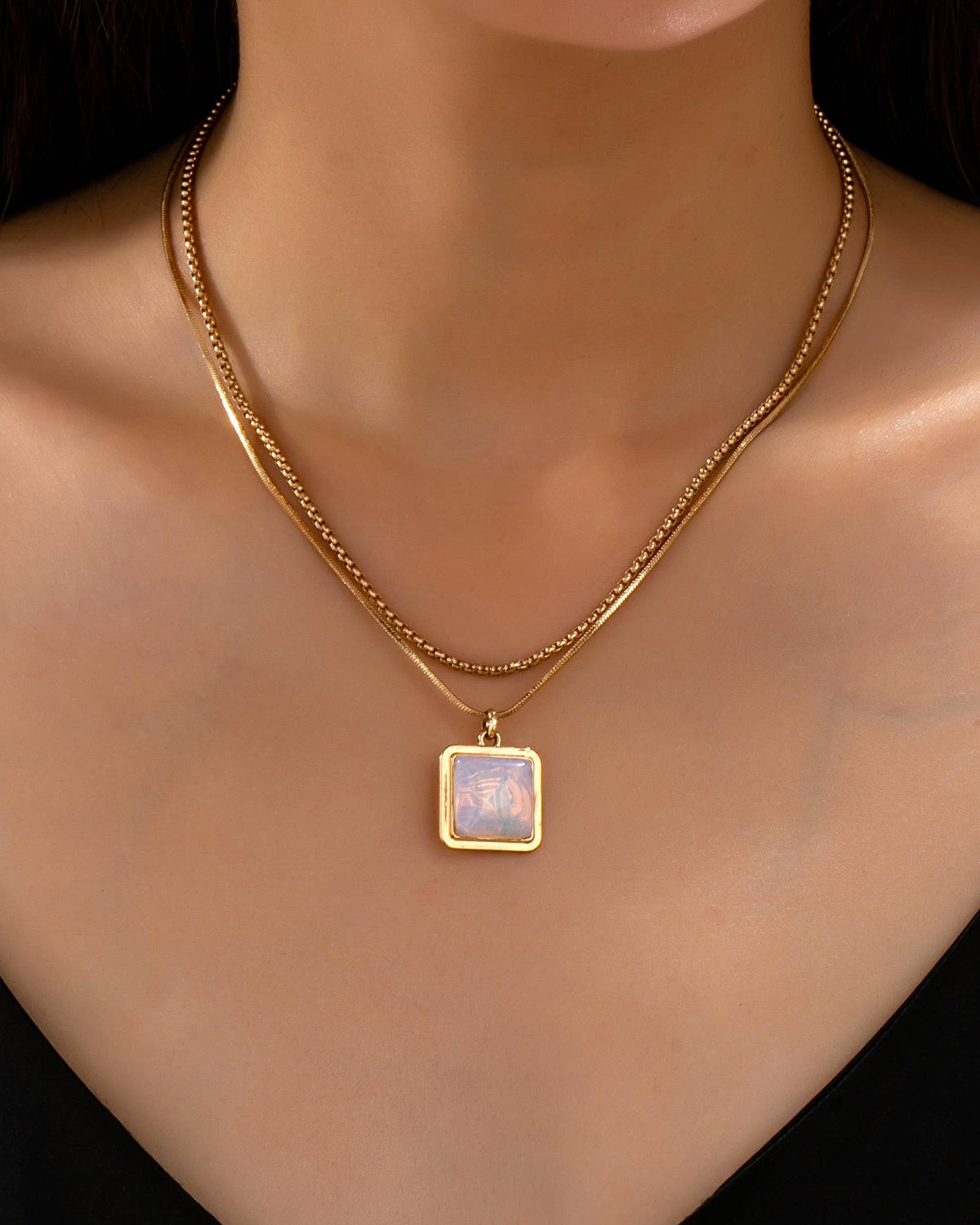 A close-up of a model's neck and chest wearing a gold double-strand chain necklace with a square pendant featuring a white opal iridescent inlay.