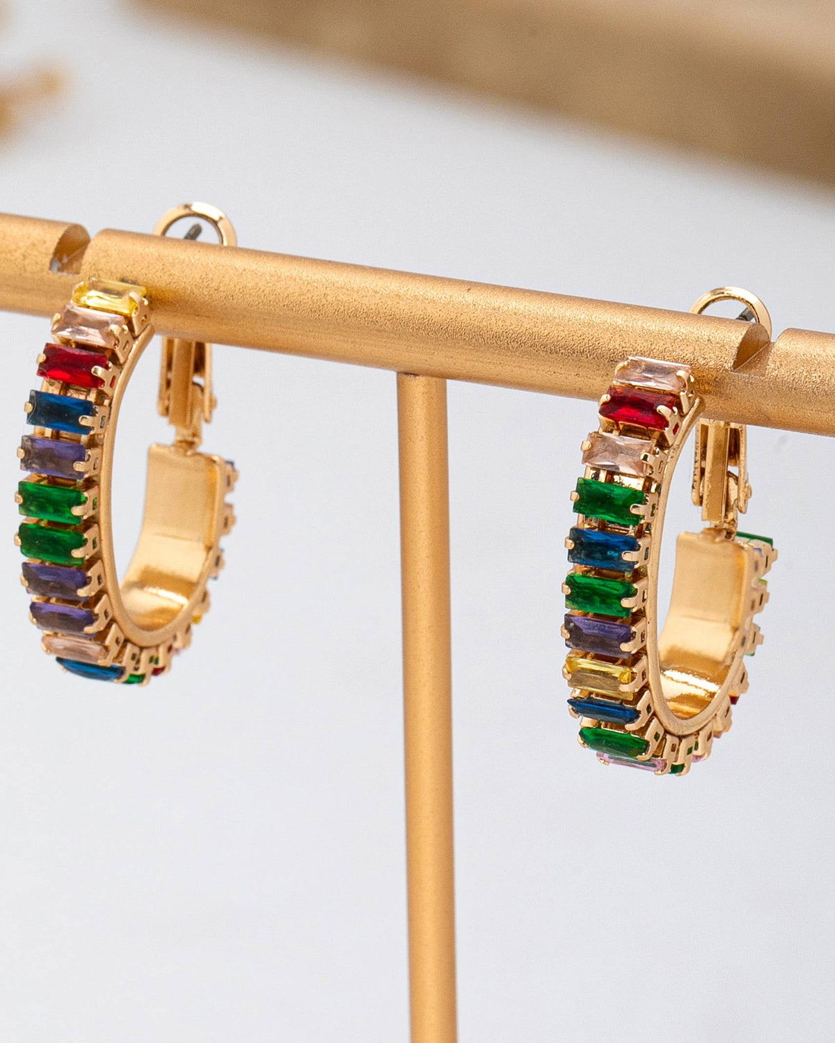 A pair of gold hoop earrings with rectangular multicolor gemstones, displayed on a gold stand.