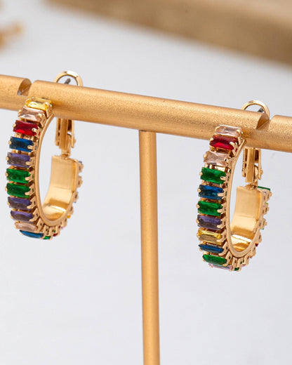 A pair of gold hoop earrings with rectangular multicolor gemstones, displayed on a gold stand.