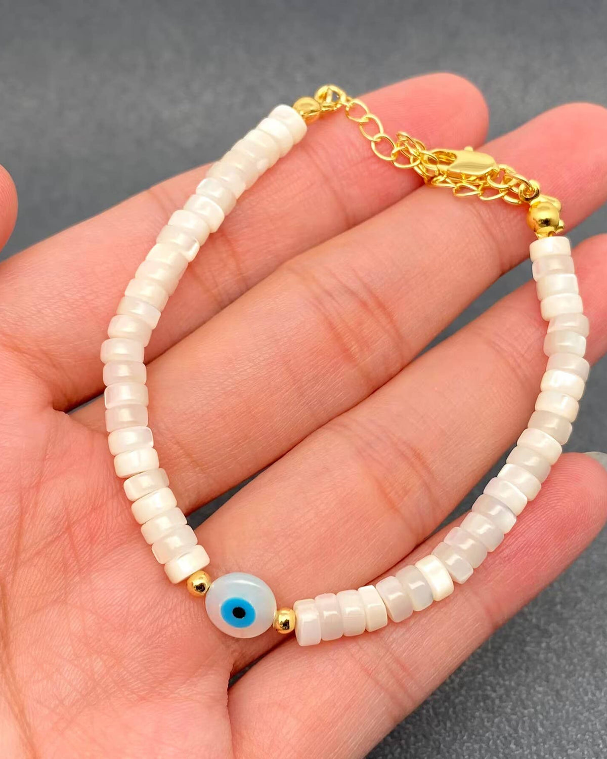 A beaded bracelet with white cylindrical beads, gold-toned metal accents, and a central blue-and-white evil eye charm, shown resting on a person's hand against a gray background.