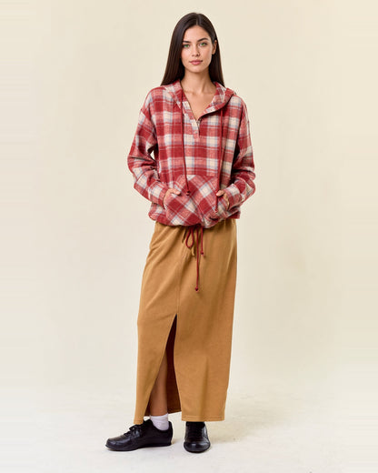 Full-length model wearing cranberry plaid flannel hoodie with hands in the kangaroo pocket and a tan skirt.