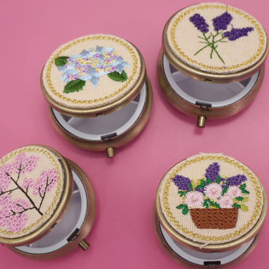 Four round embroidered compact mirrors with gold-tone metal frames, shown open on a solid pink background. Each compact lid features a different floral or botanical embroidery: purple lavender, blue hydrangea, pink cherry blossoms, and a basket of pink and purple flowers.