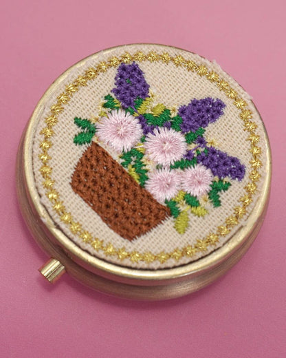 Round embroidered pill box with flowers on a pink background.