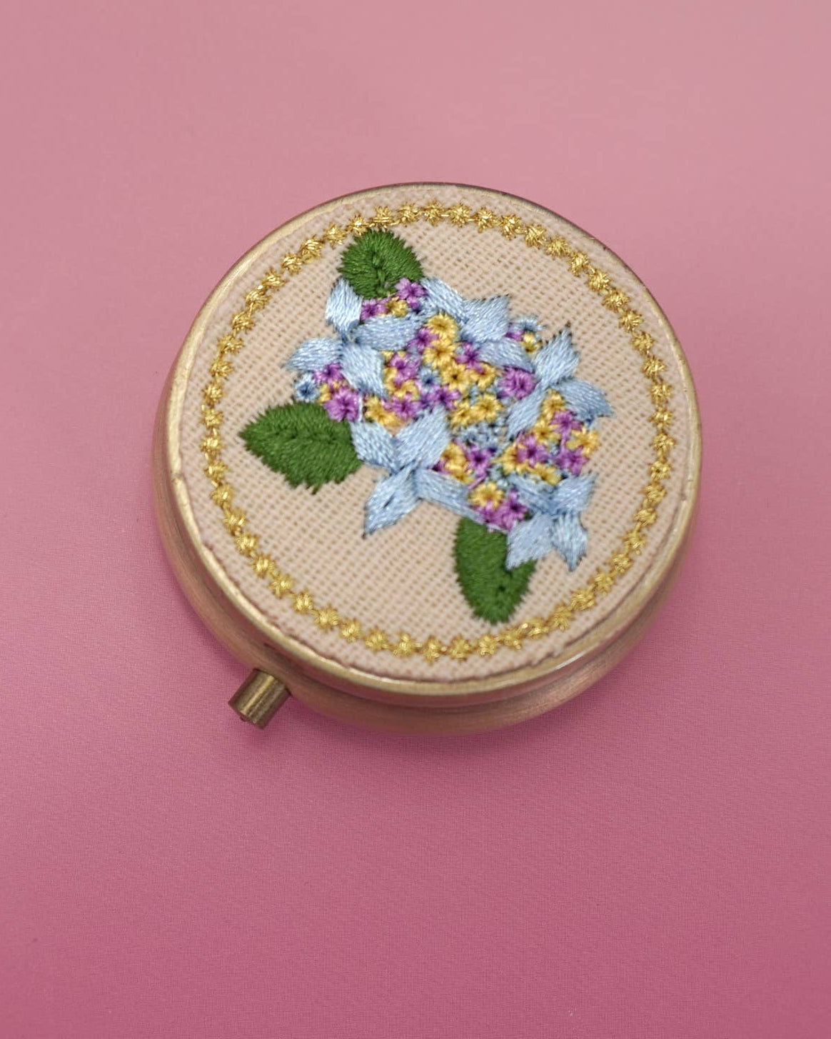 A round embroidered compact pill case with a floral design, featuring light blue and purple flowers with green leaves on a beige fabric background, set against a solid pink backdrop.