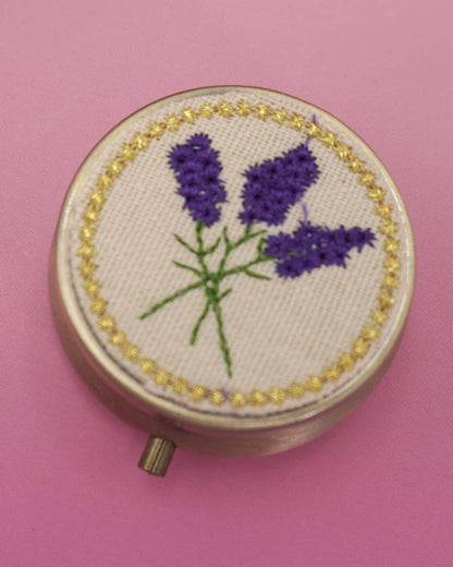 Embroidered compact pill case with lavender design on a pink background.