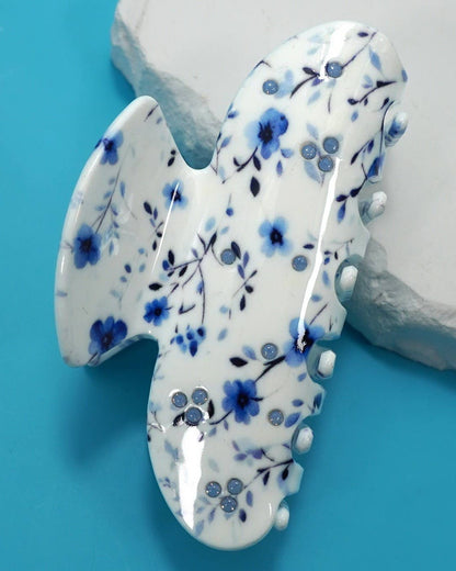 A glossy white hair claw clip decorated with a blue floral pattern and small rhinestone accents, displayed on a bright blue background with a white stone prop.