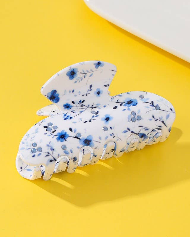 A white claw-style hair clip decorated with a blue floral pattern, resting on a bright yellow background.