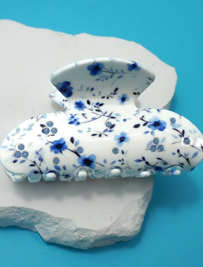 A white hair claw clip decorated with a blue floral pattern, displayed on a white stone prop against a bright blue background.