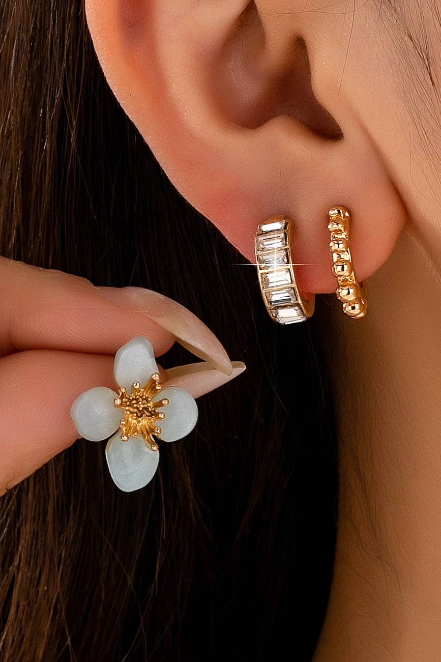 Close-up of a person's ear wearing two small gold hoop earrings, one crystal and one textured, with a hand holding a green flower-shaped earring with a gold center.