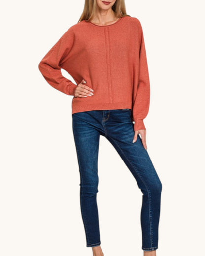 A woman wearing a rust-orange knit sweater with long batwing sleeves and dark blue skinny jeans, standing against a plain off-white background.