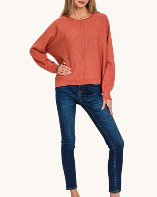 A woman wearing a rust-orange knit sweater with long batwing sleeves and dark blue skinny jeans, standing against a plain off-white background.