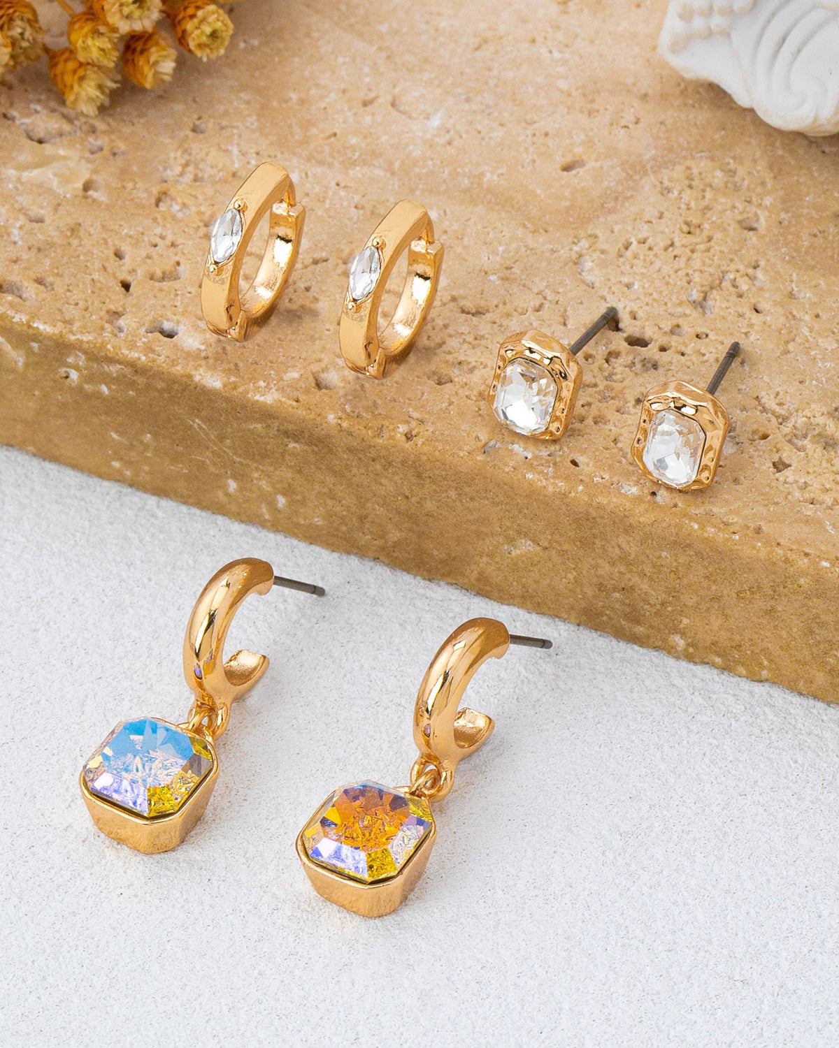 A trio set of gold-toned earrings displayed on a textured stone slab and white surface, including two small hoop earrings with crystals, two stud earrings with crystals, and two larger gold huggie drop earrings with iridescent sparkling crystal. 