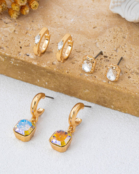 A trio set of gold-toned earrings displayed on a textured stone slab and white surface, including two small hoop earrings with crystals, two stud earrings with crystals, and two larger gold huggie drop earrings with iridescent sparkling crystal. 