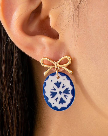 A close-up of a person's ear wearing a dangling earring with a gold bow-shaped top and an oval blue-and-white patterned charm.