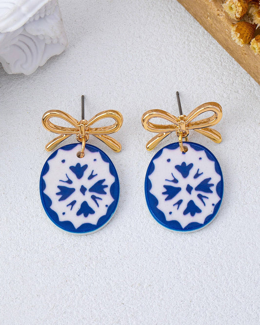 Pair of earrings with blue and white floral designs and gold bows on a light background
