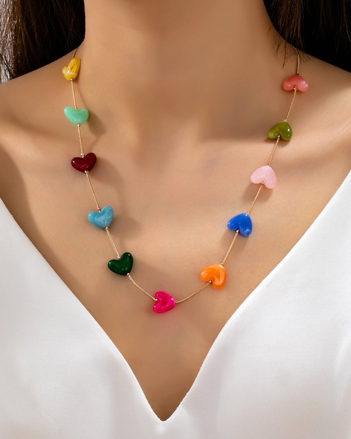 A close-up of a model wearing a delicate gold chain necklace adorned with colorful heart-shaped beads in yellow, green, red, pink, blue, orange, and dark green, styled with a white V-neck blouse.