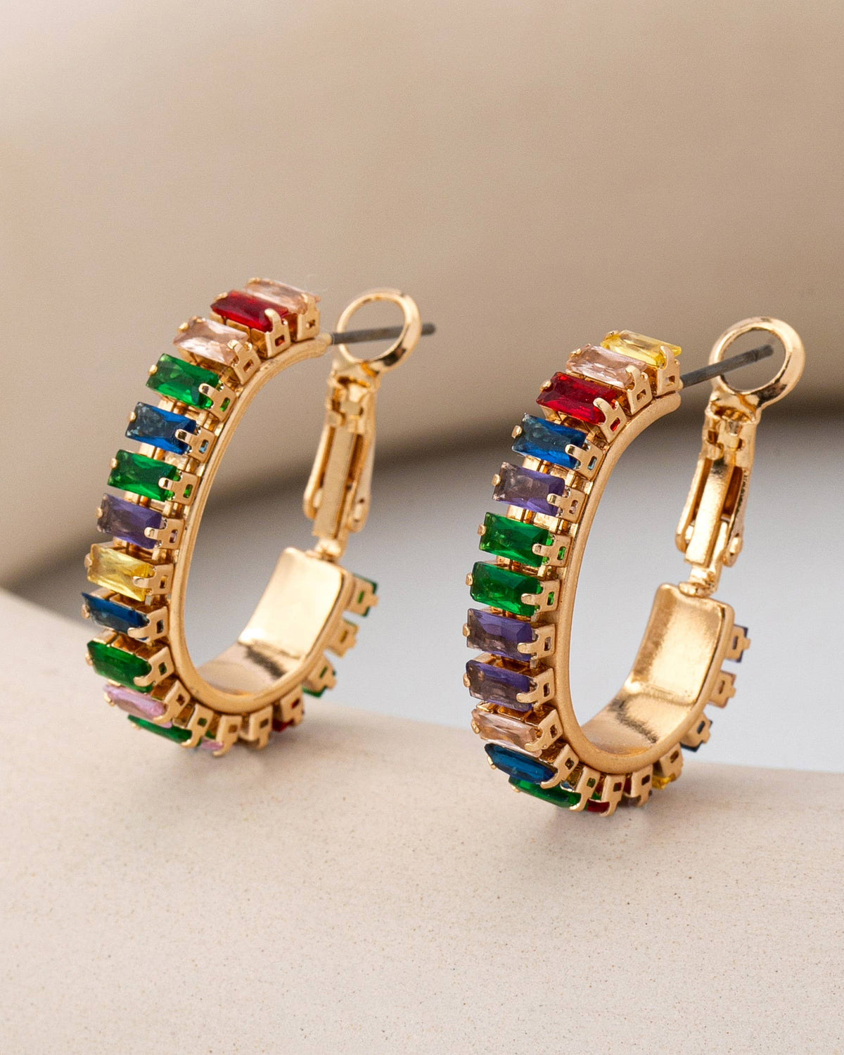A pair of gold hoop earrings decorated with colorful rectangular gemstones in red, green, blue, purple, and yellow, displayed on a neutral beige surface.