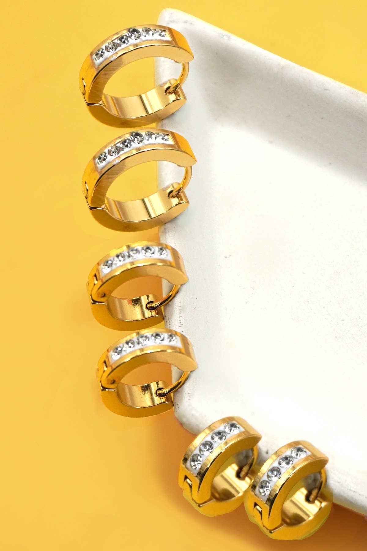 3 pairs of gold  huggie hoop earrings with clear gemstones on a yellow background.