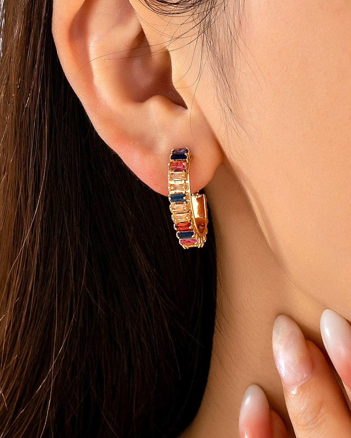 A close-up of a model's ear wearing a gold huggie hoop earring adorned with alternating rectangular jewel toned crystals.