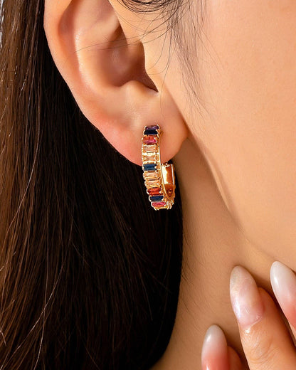A close-up of a model's ear wearing a gold huggie hoop earring adorned with alternating rectangular jewel toned crystals.