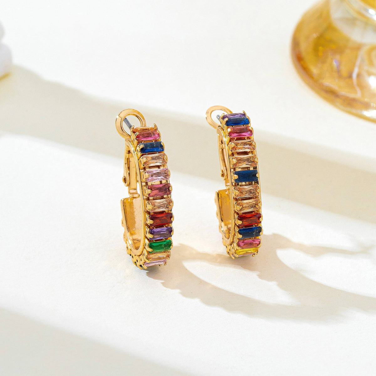 A pair of gold hoop earrings decorated with colorful rectangular gemstones in pink, blue, red, green, and purple, displayed on a clean white surface.