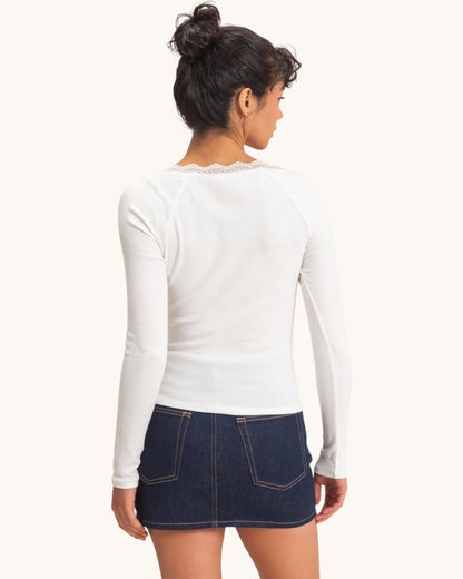 A woman shown from the back wearing a fitted white long-sleeve top with delicate lace trim at the neckline and a dark blue denim mini skirt, against a plain off-white studio background.