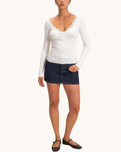 A woman modeling a white long-sleeve top with a scalloped V-neckline and gathered bust, paired with a dark denim mini skirt and black flats, against a plain off-white background.