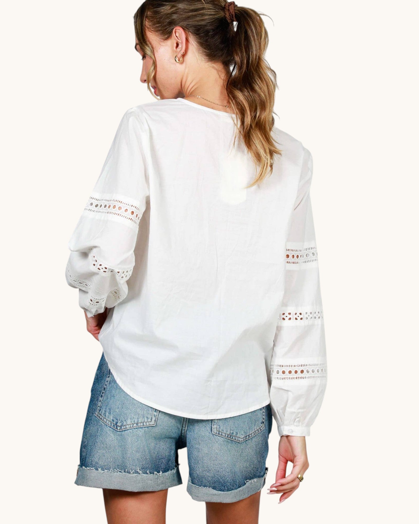 A woman shown from the back wearing a white long-sleeve blouse with eyelet lace detailing on the sleeves and denim shorts, against a plain off-white background.