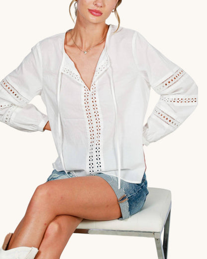 A woman wearing a white long-sleeve eyelet blouse with drawstrings and denim shorts, seated on a white stool against a light beige studio background.