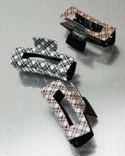 Three plaid hair claw clips in black, brown, and tan tones, arranged diagonally on a brushed metal surface.