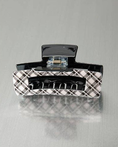 A hair claw clip with a black glossy top and bottom and a black-and-white diagonal plaid pattern on the sides, shown on a reflective gray surface.