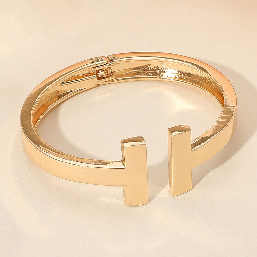 Gold bracelet with a 'T' design on a beige background.