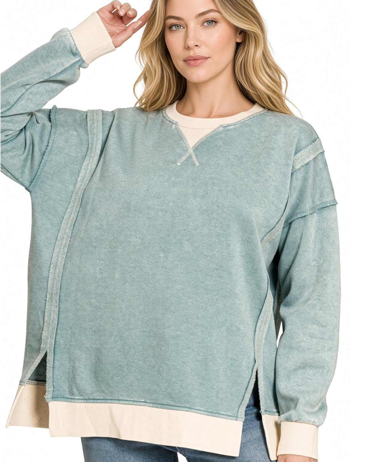 A woman modeling a long-sleeve, oversized ash-blue sweatshirt with contrasting cream ribbed trim at the crew neck, cuffs, and hem, paired with blue jeans against a plain white background.