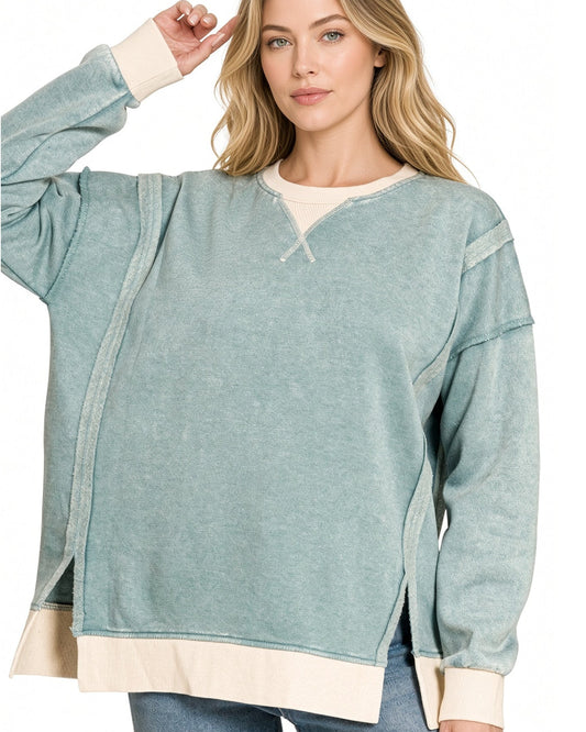 A woman modeling a long-sleeve, oversized ash-blue sweatshirt with contrasting cream ribbed trim at the crew neck, cuffs, and hem, paired with blue jeans against a plain white background.